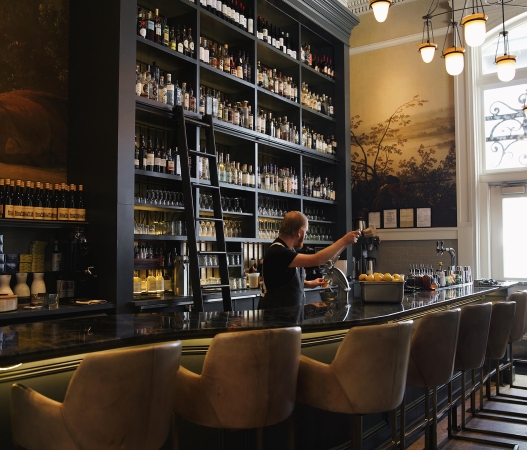 Tall shelving of assorted liquors behind the bar at Ultreia at Union Station in Denver, Colorado. A bar tender prepares drinks.
